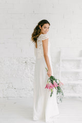 Novia de pie, sonriendo a cámara sobre el hombro. Lleva un vestido espalda descubierta para novia boho. En las manos tiene un ramo silvestre, Al lado hay una escalera.