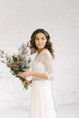 Modelo de perfil vestida de novia. La sobrecapa que lleva le permite llevar los brazos tapados con encaje gracias al abrigo con cola