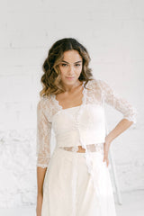 Modelo con el brazo en jarra llevando un traje de novia que consiste en unos pantalones, un crop top y un abrigo de encaje ivory