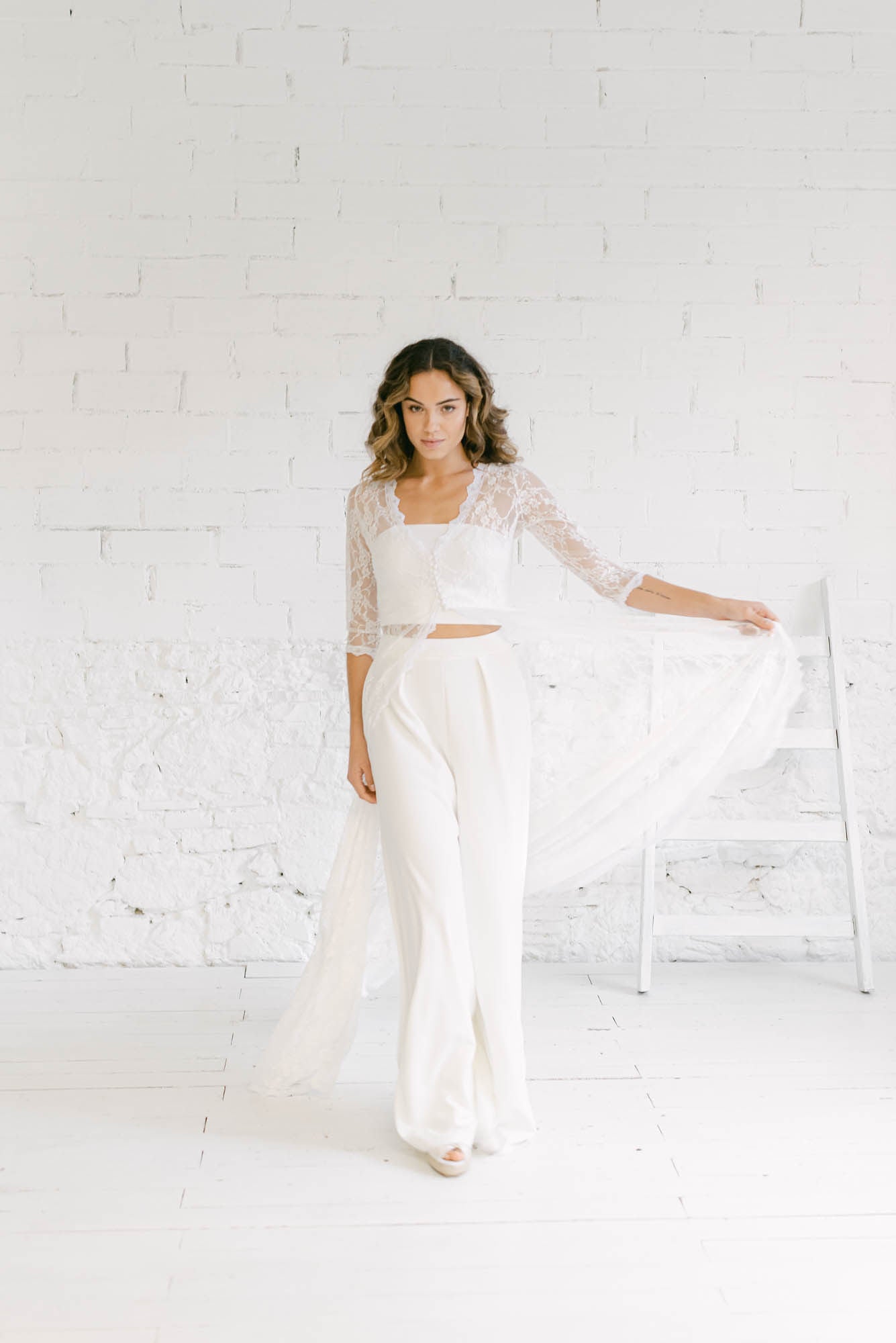 Modelo vestida de novia caminando hacia cámara. Lleva un traje dos piezas con un abrigo de encaje que mueve con los brazos