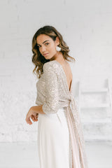 Modelo chica con vestido de novia dos piezas compuesto por pantalón palazzo blanco ancho y top de manga tres cuartos en tela de lentejuelas plateadas con preciosa espalda abierta con dos lazos anudados.