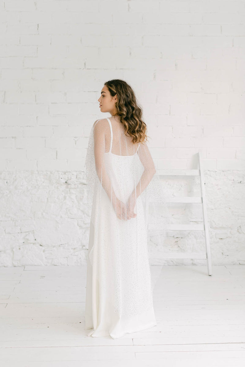 Modelo de espaldas con un vestido brillante novia de espalda baja y escote en V. Encima lleva como accesorio ideal una capa de tul brilli.