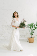 Modelo de perfil y caminando. Lleva unas flores en los brazos. Detrás hay una planta. Combina un vestido lencero con un abrigo de encaje de ivory con cola