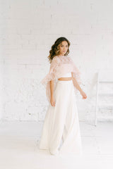 Modelo vestida como novia en perfil. Con una pierna delante de la otra, se puede ver el ancho del pantalón palazzo, súper elegante. También, el tube top y una caja con mucho vuelo de encaje rosa cuarzo. Es perfecto como vestido novia civil