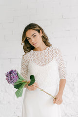 Modelo con un vestido novia rustico con crop top. Está sonriendo a cámara con una flor en las manos. El crop top está hecho un tul bordado con pequeñas flores