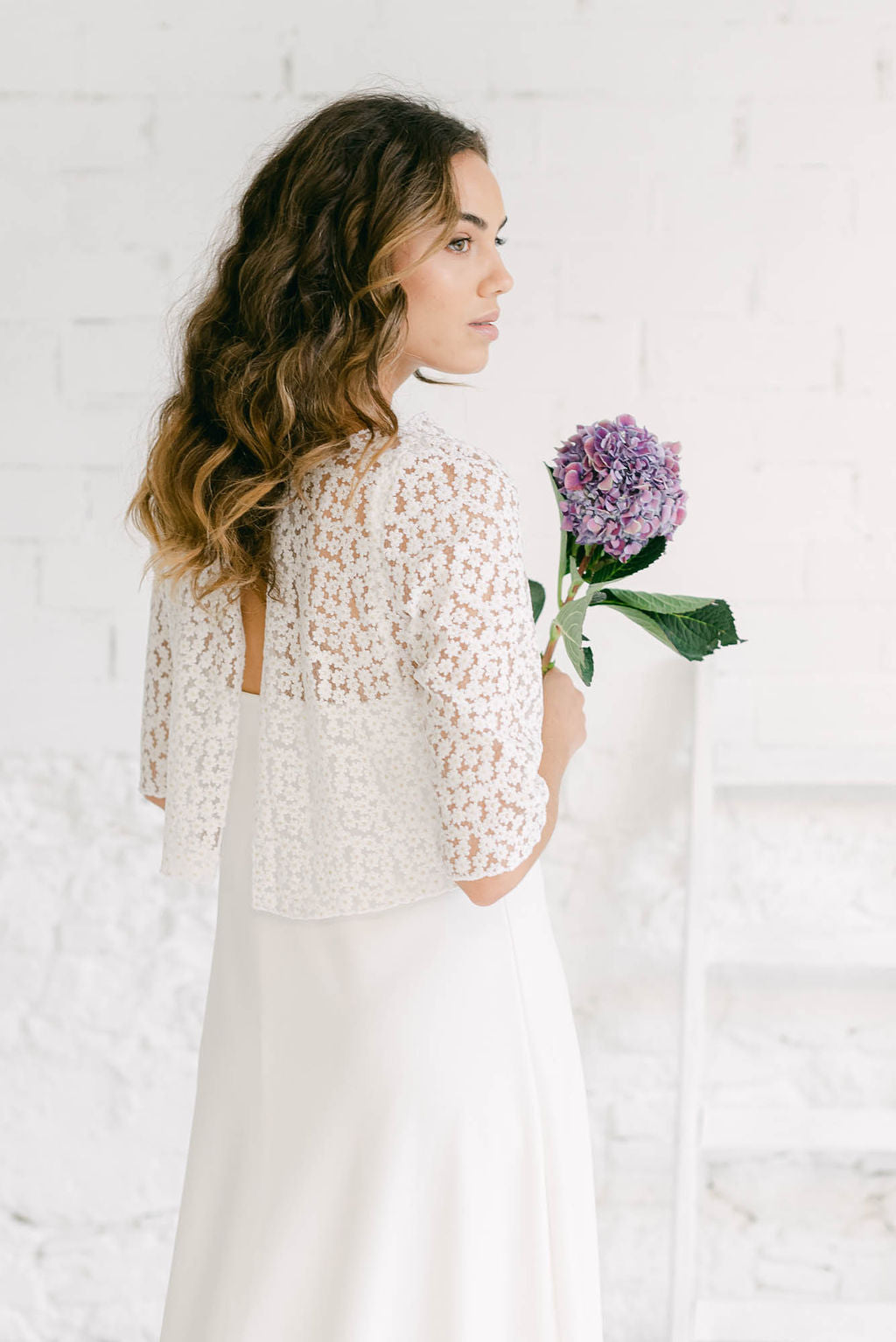 Modelo de perfil con una flor en la mano. Lleva puesto un vestido rústico de novia. Combina un modelo lencero con un crop top de mangas tres cuartos.