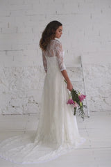 Video de modelo de espaldas a cámara con abrigo con cola larga de encaje espectacular. La modelo lleva ramo de flores rosa en la mano.