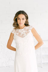 Modelo chica mirando a cámara con vestio de novia blanco de tirantas y crop top de flores blancas bordadas de mangas cortas por encima con espalda abierta.