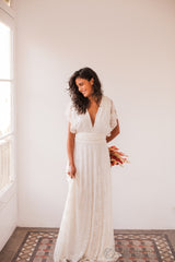 Una mujer sonríe mientras lleva puesto un vestido de novia de encaje marfil con escote en V, sosteniendo un elegante ramo de flores