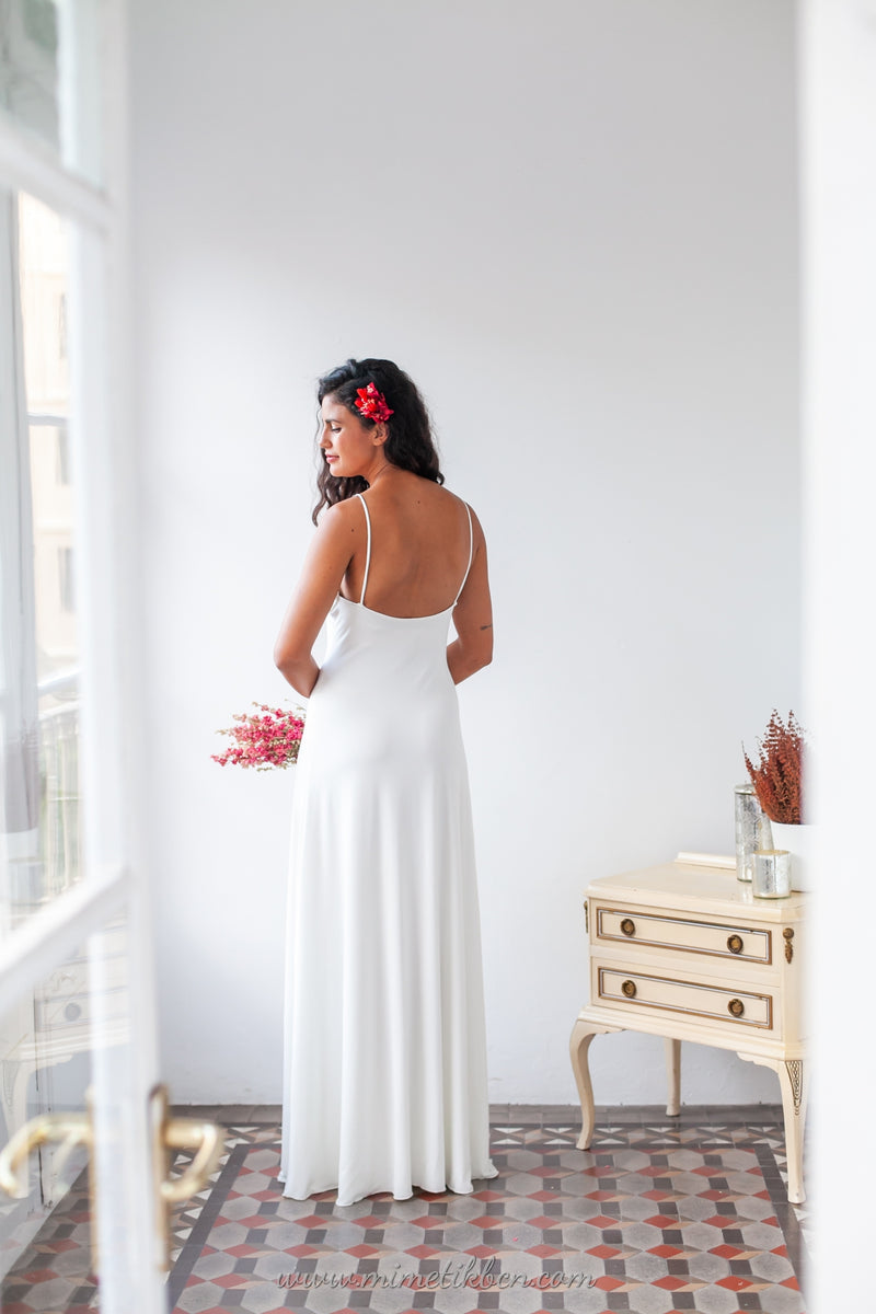 El vestido de novia blanco Marie, con su diseño atemporal y tirantes, se muestra desde atrás mientras la novia sostiene un encantador ramo de flores