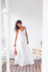 Detalle del vestido de novia blanco Marie, largo de tirantes, llevado por una modelo con un ramo de flores en una mano