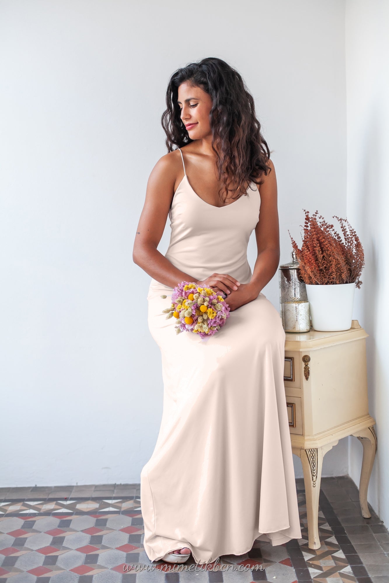 Una mujer se sienta elegantemente en una pequeña mesa mientras luce un vestido de novia color champagne con tirantes