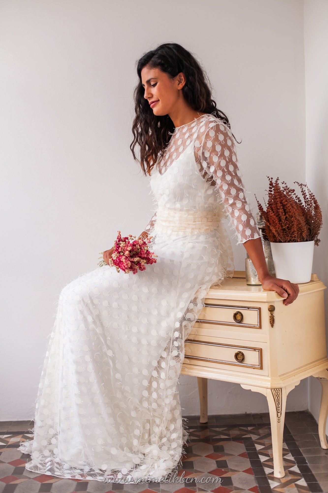 En el ambiente acogedor de un atelier, la novia se sienta con gracia luciendo un sobrevestido de tul sobre su deslumbrante vestido de novia blanco de Marie