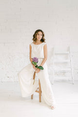 Modelo sentada en banquito de madera con traje de novia dos piezas formado por pantalon palazzo blanco y crop top blanco de color blanco con espalda abierta en V inversa.