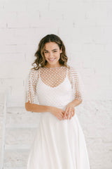 Modelo frente a cámara y sonriendo con brazos en la cintura. La chica lleva un vestido de novia dos piezas con falda blanca y top con tela de encaje con escote en la espalda y florecitas blancas.