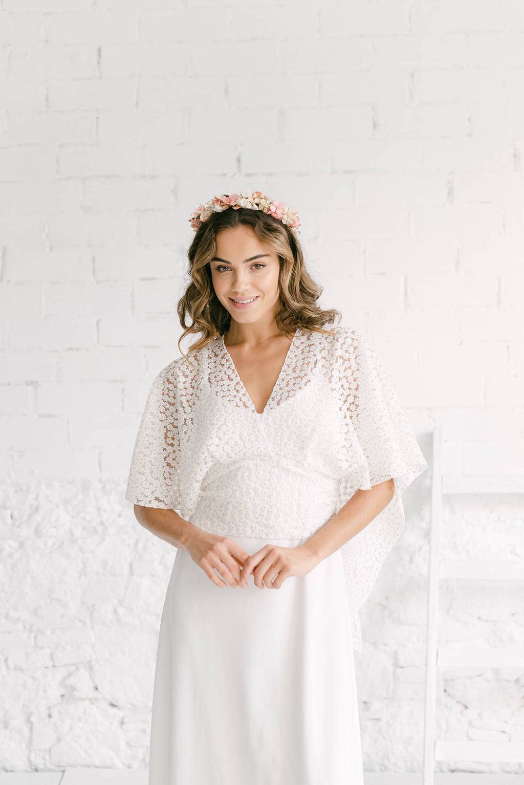 Modelo sonriendo a cámara con diadema de flores corales en la cabeza. La modelo lleva el vestido Marie de tirantes blanco con la capa lilly con margaritas doradas.