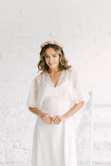 Modelo sonriendo a cámara con diadema de flores corales en la cabeza. La modelo lleva el vestido Marie de tirantes blanco con la capa lilly con margaritas doradas.