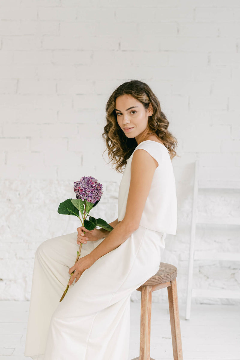 Modelo sentada en banco de madera con ramo de flores lilas en la mano. La chica lleva un vestido de novia dos piezas de pantalón palazzo y crop top blanco con espalda abierta.