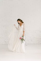 Chica modelo con vestido de novia dos piezas con abrigo por encima con cola larga de encaje moviendo. La chica mueve la capa del abrigo con la mano derecha.