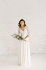 Chica modelo frente a cámara con un vestido de novia dos piezas formado por falda de crepe elastica con crop top de tirantes blanco y abrigo de encaje con cola por encima.