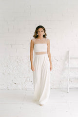 Modelo chica frente a cámara con traje de novia dos piezas formado por pantalón palazzo blanco de vuelo y crop top blanco en tela de tul de florecitas blancas con top palabra de honor blanco por debajo.
