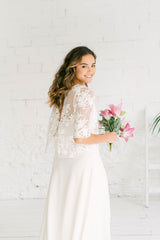 Modelo chica de semi-espaldas y sonriendo a la cámara, con ramo de flores en la mano. Lleva puesto un vestido largo de tirantasblanco y crop top por encima con espalda abierta en pico en tela de tul transparente de encaje blanco.