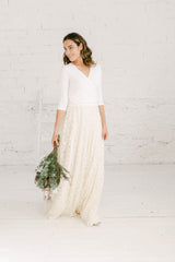 Modelo chica mirando hacia la derecha con ramo de flores en la mano. La modelo lleva un vestido de novia dos piezas con falda color crema y top manga tres cuartos blanca,