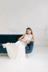 Modelo chica sentada en sofa azúl. La modelo lleva un vestido dos piezas de novia compuesto por falda blanca + tube top palabra de honor blanco y crop top de flores con espalda abierta.