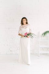 Modelo mirando a camara sujetando ramo de flores rosas en la mano con vestido de novia blanco de tirantas y escote de pico y crop top de manga francesa en tela de tul de flores blancas.