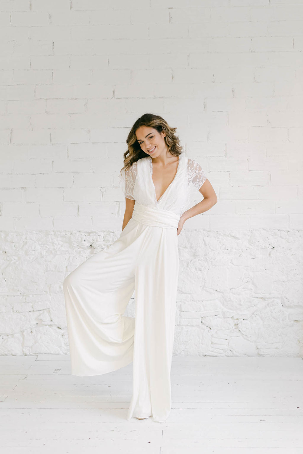 Modelo chica sonriendo a cámara con pierna derecha levantada y brazo izquierdo en la cintura. La modelo lleva un vestido de novia original con mono pantalon blanco Coco y straps en el escote haciendo unas mangas en tela de encaje blanco.