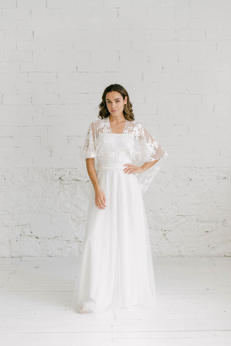 Modelo de pie luciendo un vestido de novia civil con una capa de tul para cubrir sus brazos