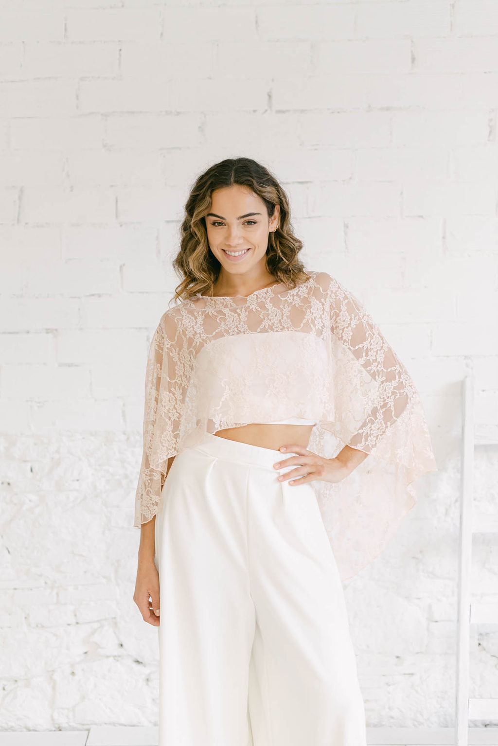 Modelo sonriendo mientras muestra una preciosa capa de novia de encaje rosa cuarzo que le queda muy sensual y elegante