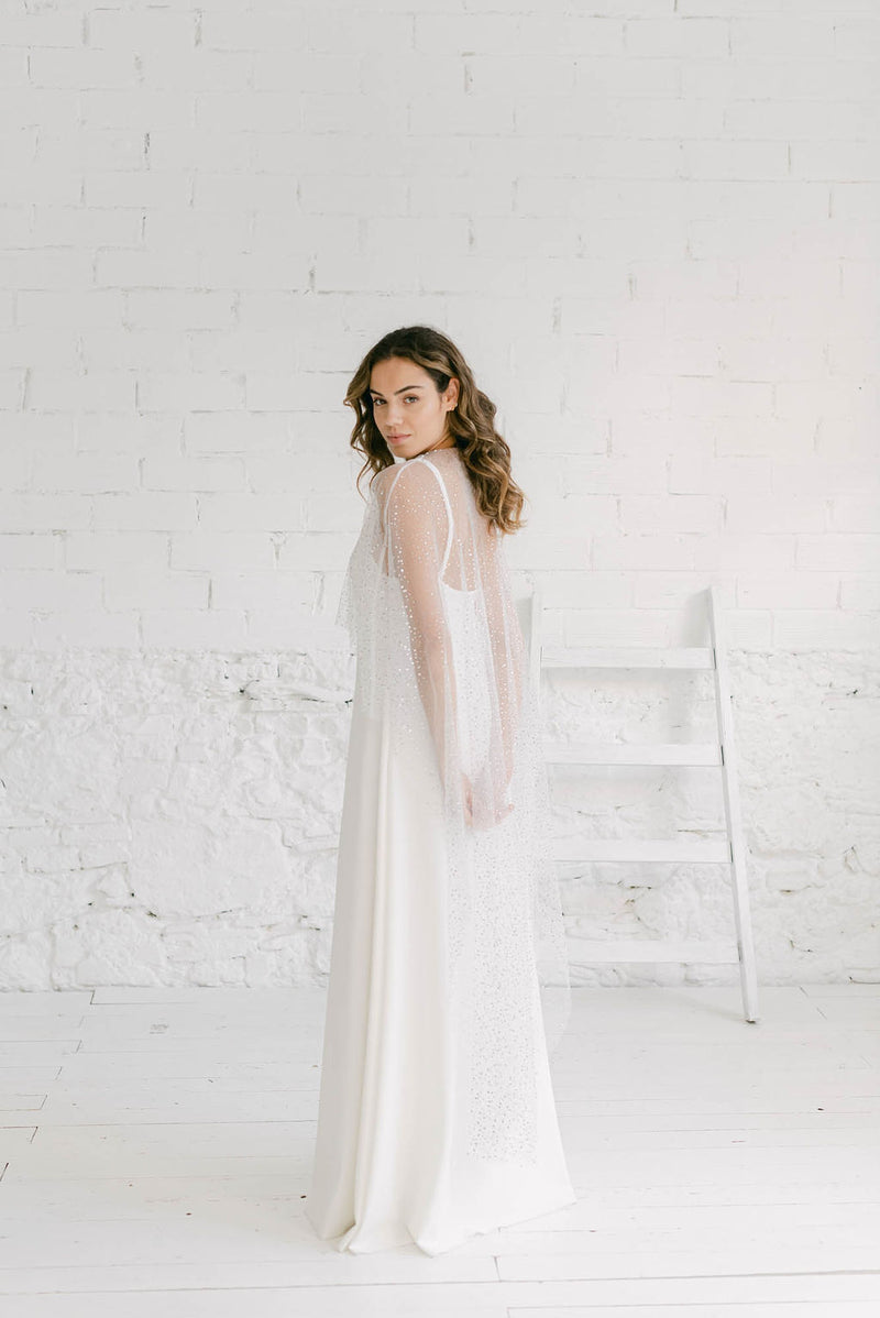 Modelo de perfil delante de una pared blanca llevando una combinación mágica: un vestido sencillo con una capa de tul brilli brilli. Es un vestido brillante de novia perfecto.