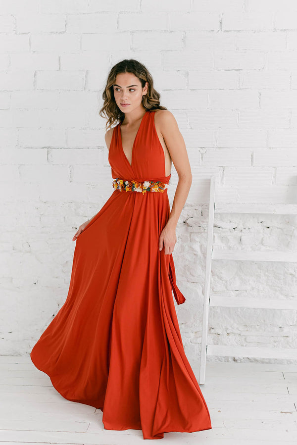 Modelo con vestido de dama de honor color terra mirando hacia la derecha. Vestido con escote en V y cinturón de flores en tonalidades naranjas y blancos.
