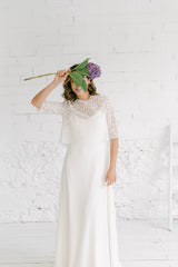 Modelo de pie tapándose la cara con una flor. Luce un vestido de novia rústico perfecto para bodas sencillas, alternativas o bohemias