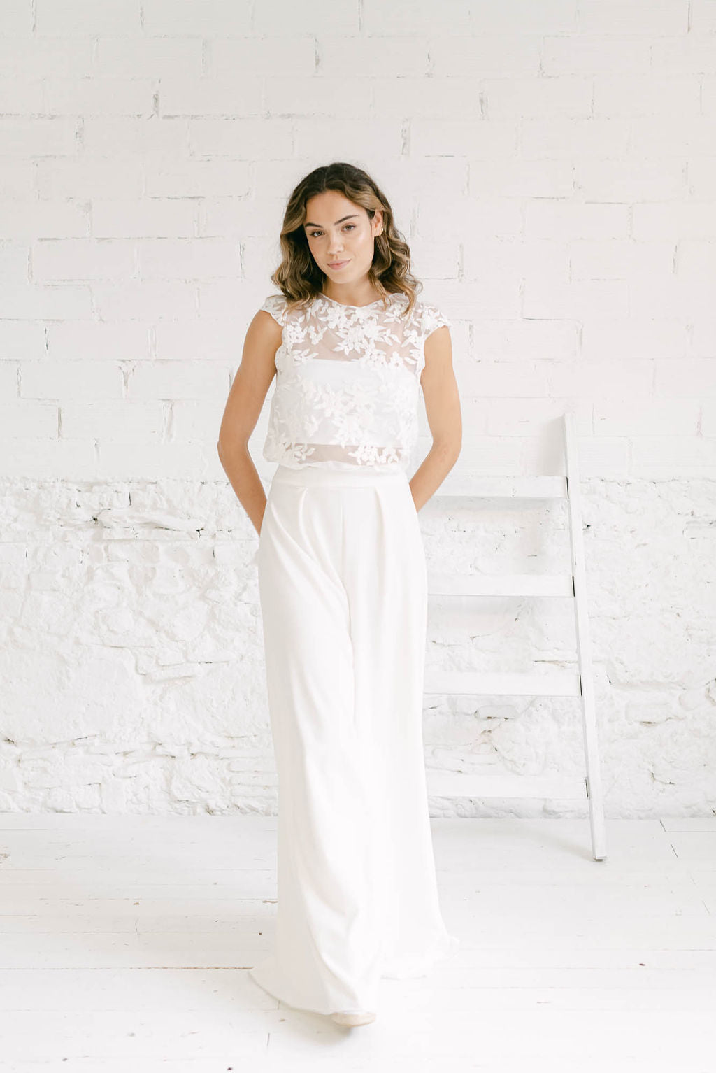 Modelo de cuerpo entero sonriendo de medio lado. Lleva puesto un vestido dos piezas con pantalón, tube top y crop top de tul de flores