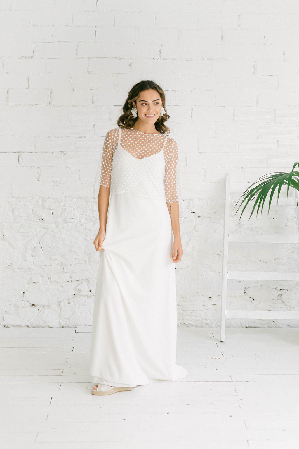 Novia de pie y sonriendo a cámara con su vestido con mangas estilo lencero. Las mangas son tres cuartos. La espalda queda abierta.