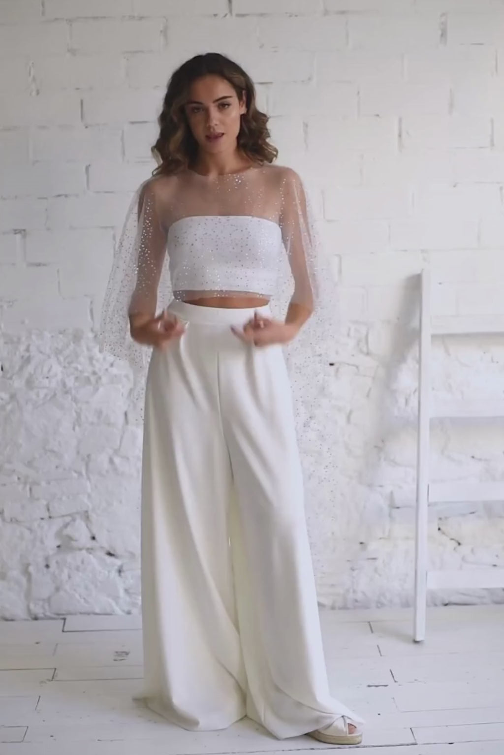 Video de modelo andando a camara con capa maxi en tela de tul de brillantes, top palabra de honor blanco y pantalon palazzo blanco.