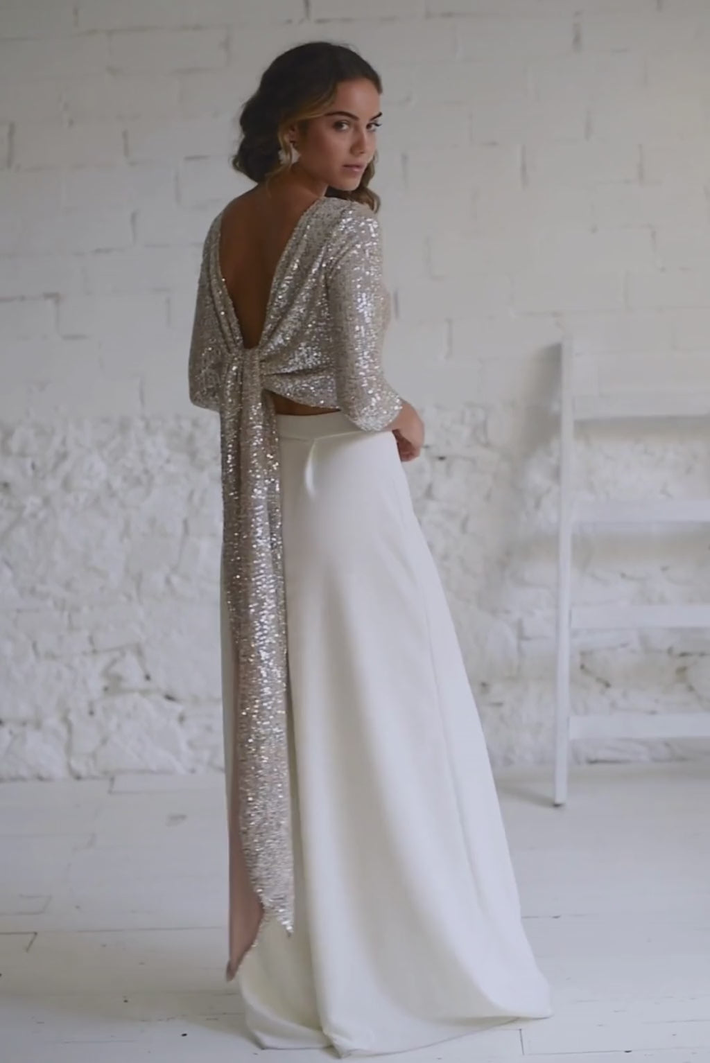 Video de modelo de espaldas con traje de novia dos piezas pantalon palazzo y top de manga larga de lentejuelas plateadas. Podemos observar la preciosa espalda abierta.