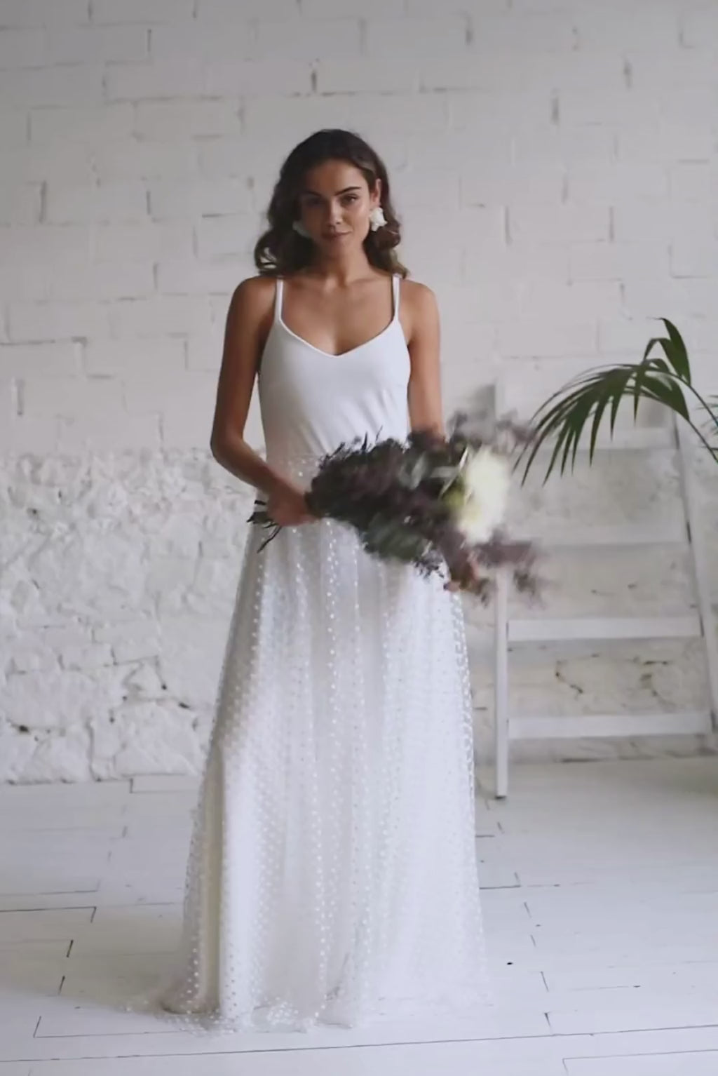 video de modelo de novia con vestido blanco liso largo con escote en V y tirantas; sobre falda en tela de tul transparente de florecitas blancas.