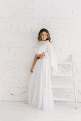 Modelo apoyada en pared blanca con brazo doblado y sonriendo hacia la izquierda. Falda de encaje blanca larga y capa rosa formato midi.