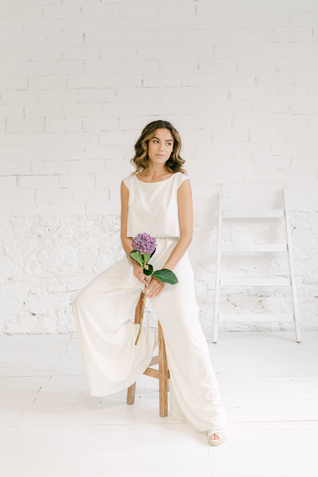 Modelo sentada en banquito de madera con traje de novia dos piezas formado por pantalon palazzo blanco y crop top blanco de color blanco con espalda abierta en V inversa.