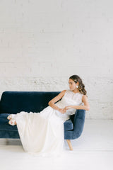 Modelo tumbada en sofá azul mirando hacia la derecha. Vestido de novia dos piezas formado por falda blanca larga y crop top en tela de tul transparente de margaritas doradas + top palabra de honor blanco.