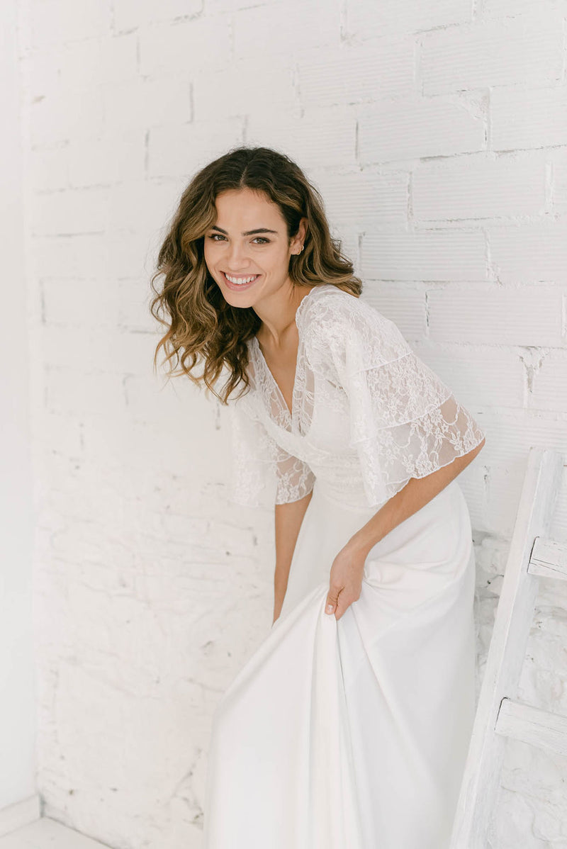 Modelo apoyada en la pared sonriendo a cámara. Sujetando el bajo de su vestido de novia blanco con top de mangas de encaje blanco.