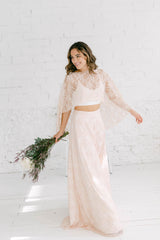 Chica vestida con traje de novia dos piezas sonriendo hacia el lado derecho y con ramo de flores lilas en la mano.