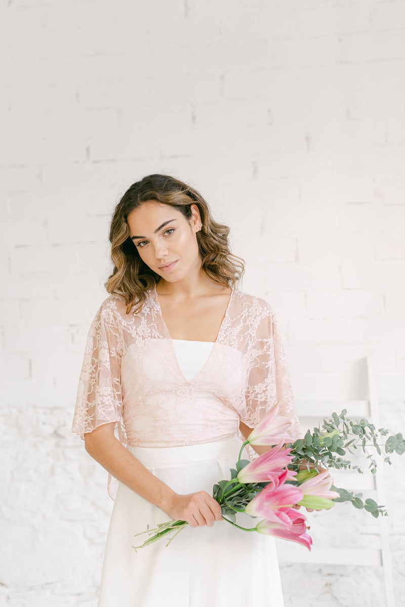 modelo mirando a cámara con ramo de flores rosas sujeto en la mano. La modelo lleva el traje de novia dos piezas con la capa Lily en color rosa cuarzo.