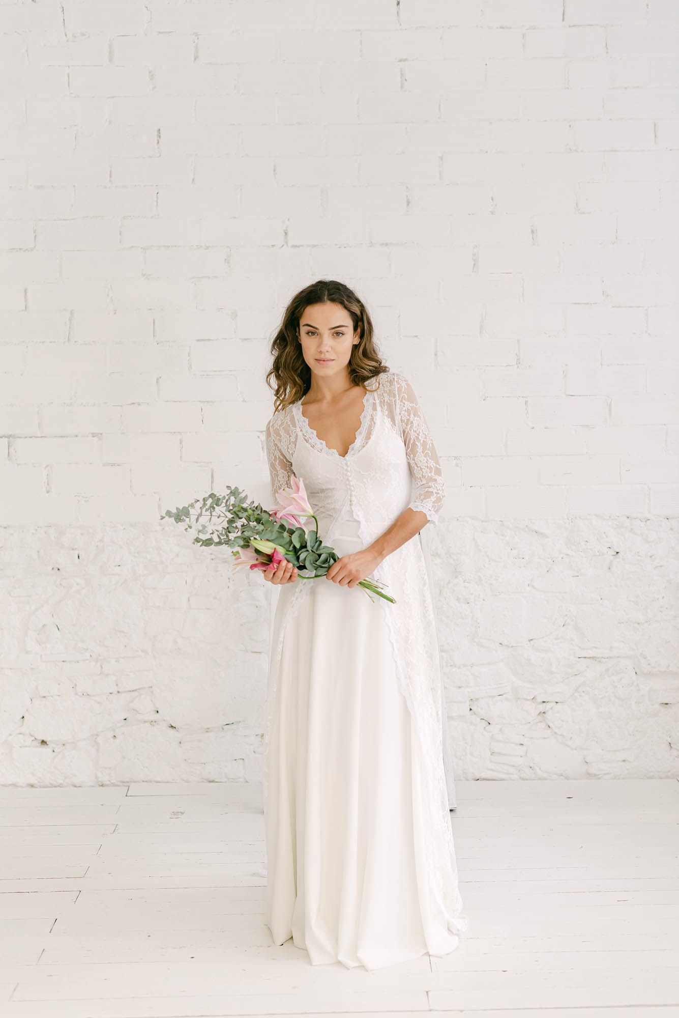 Chica modelo frente a cámara con un vestido de novia dos piezas formado por falda de crepe elastica con crop top de tirantes blanco y abrigo de encaje con cola por encima.