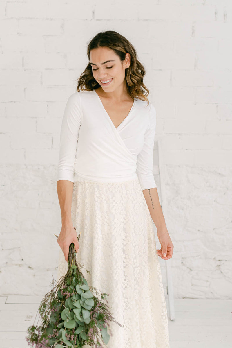 Foto más ampliada de modelo sonriendo y mirando al suelo con falda en color crema de bordado de flores y vestido blanco por debajo de manga larga.