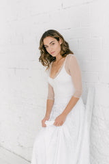 Modelo chica apoyada en una pared blanca, con vestido de novia dos piezas de encaje y transparencias.