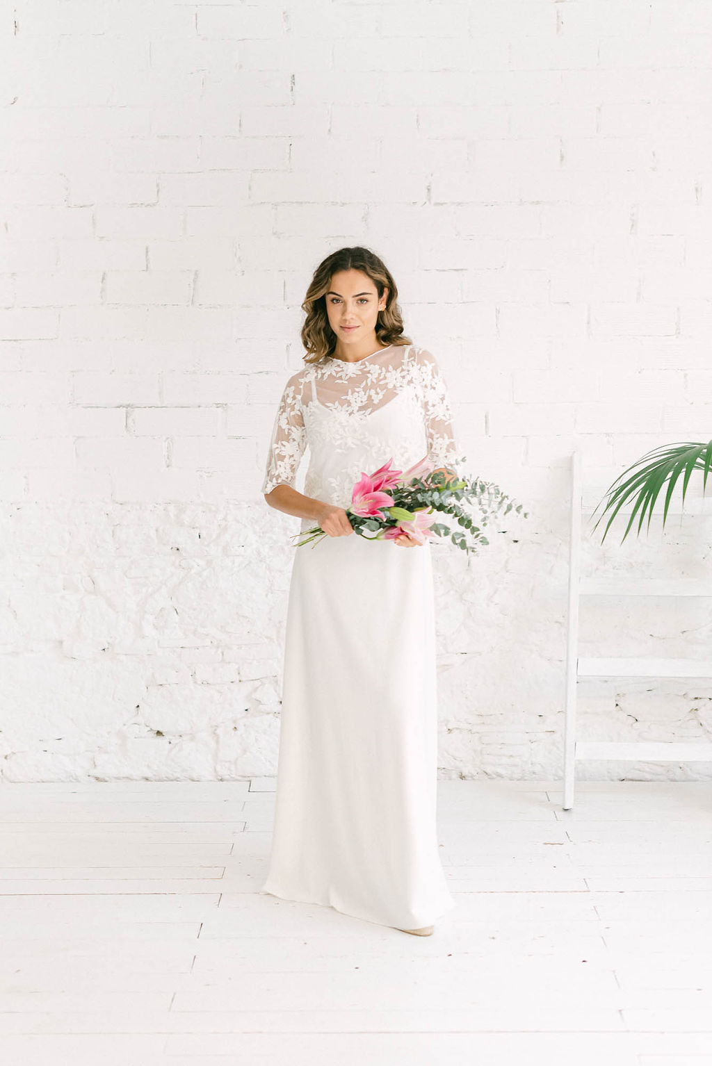 Modelo mirando a camara sujetando ramo de flores rosas en la mano con vestido de novia blanco de tirantas y escote de pico y crop top de manga francesa en tela de tul de flores blancas.